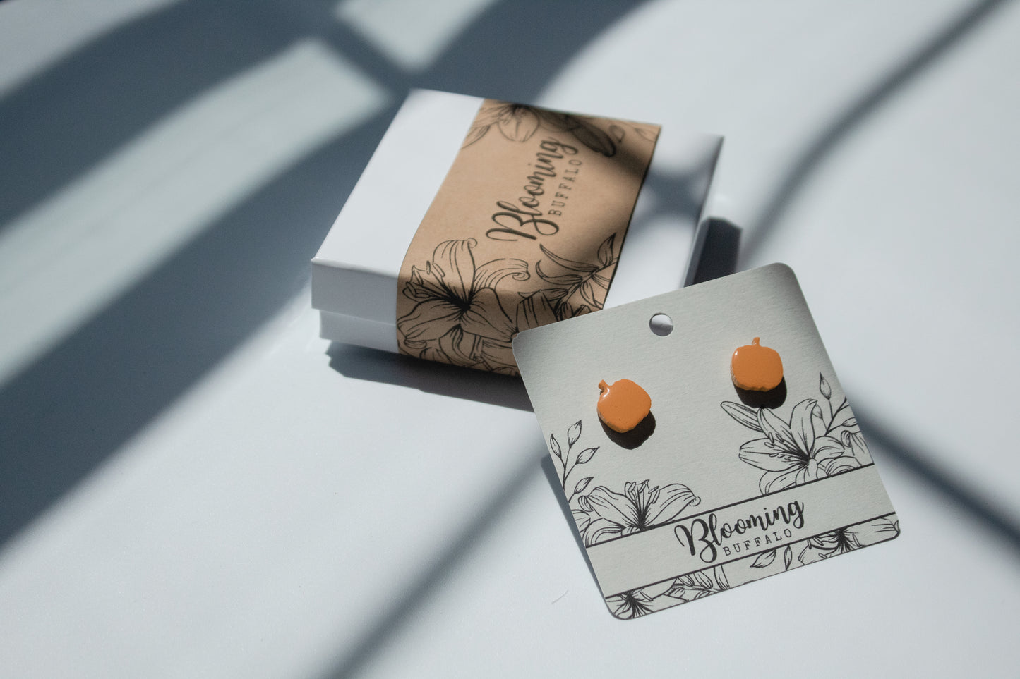 Orange pumpkin stud earrings on an earring card with the name blooming buffalo. The earring card is leaning against a white jewelry box.