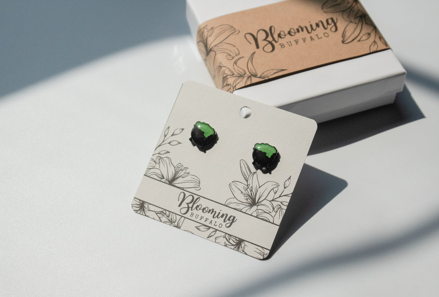 black cauldron stud earrings with vibrant green witch's brew bubbling over the side on a Blooming Buffalo branded earring card
