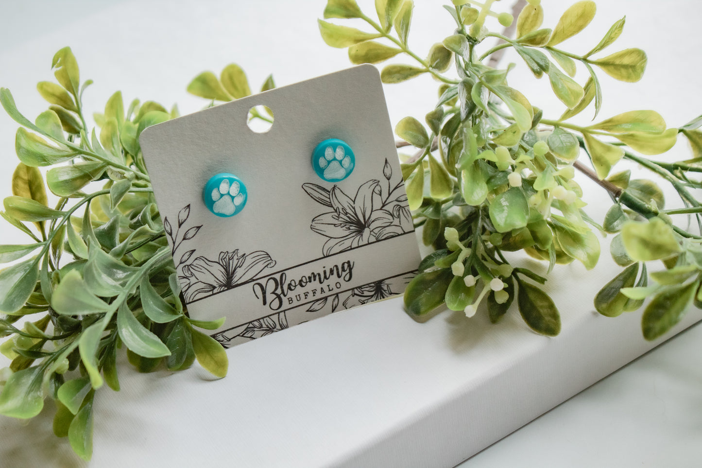 teal circle stud earrings with white paw print design on Blooming Buffalo branded earring card