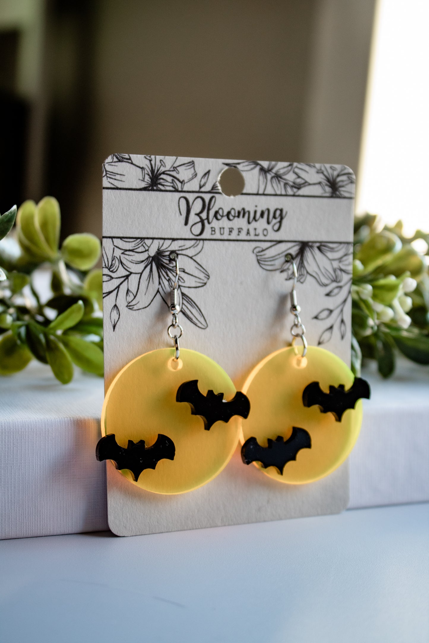 Translucent yellow full moon dangle earrings with two black bats on each earring. The earrings are on a Blooming Buffalo earring card .