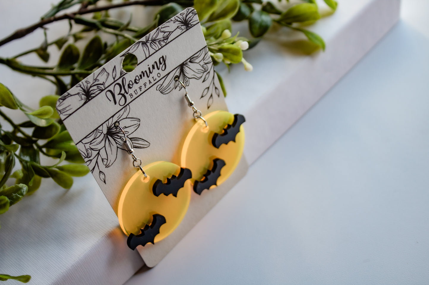 Translucent yellow full moon dangle earrings with two black bats on each earring. The earrings are on a Blooming Buffalo earring card.