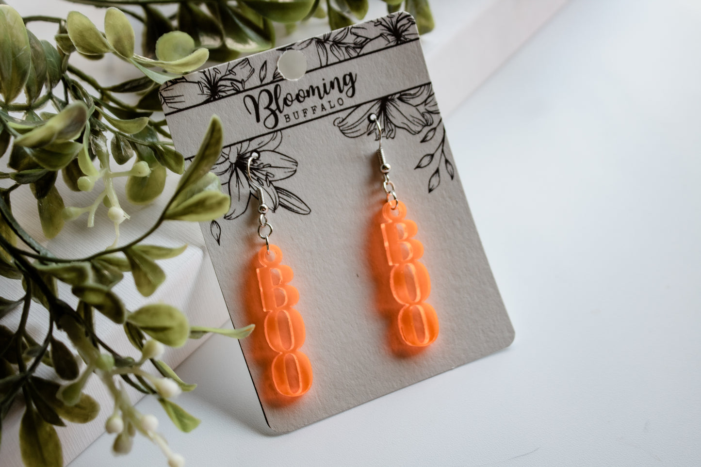 Bright fluorescent orange dangle earrings shaped like the word BOO on a Blooming Buffalo branded earring card