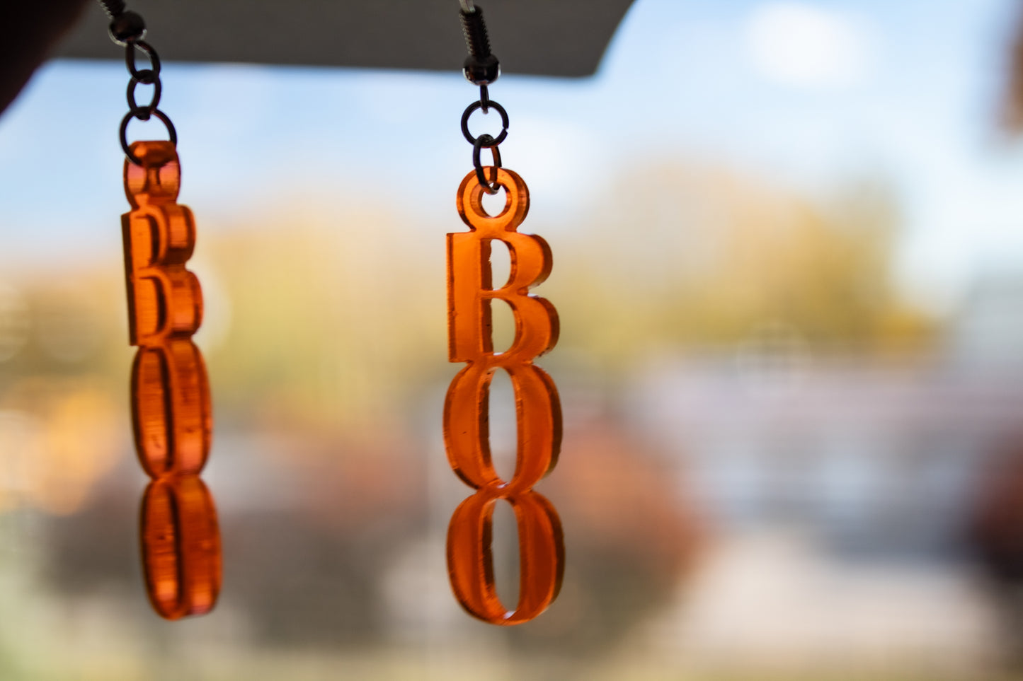 Bright fluorescent orange dangle earrings shaped like the word BOO in front of a window to show light coming through them