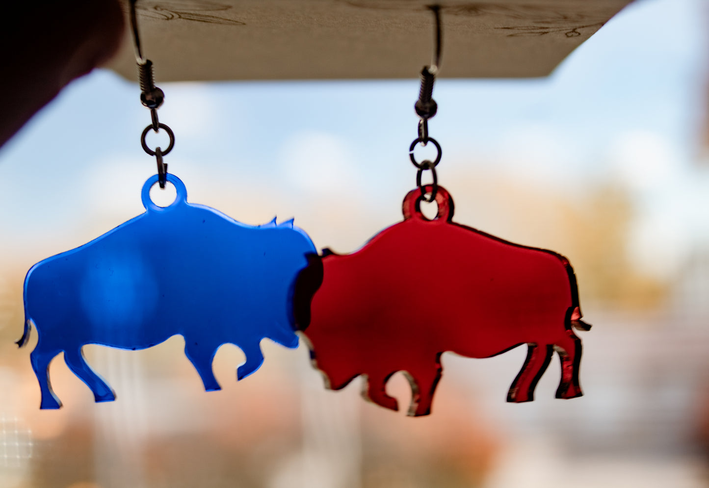 Translucent red and blue buffalo dangle earrings in front of a window showing how they look in the light