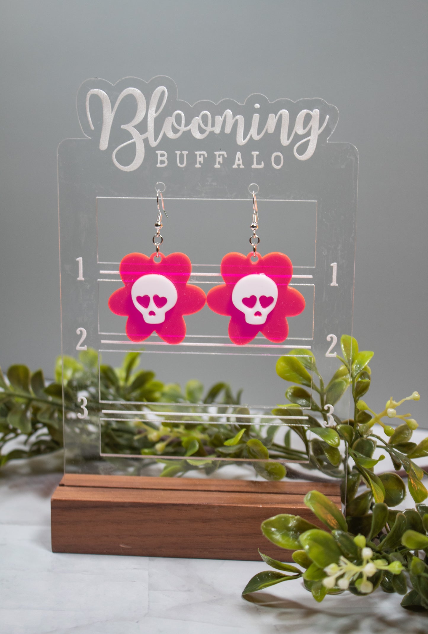 fluorescent hot pink flower dangle earrings. White skulls with heart eyes in the center. Earrings are on a Blooming Buffalo branded earring stand