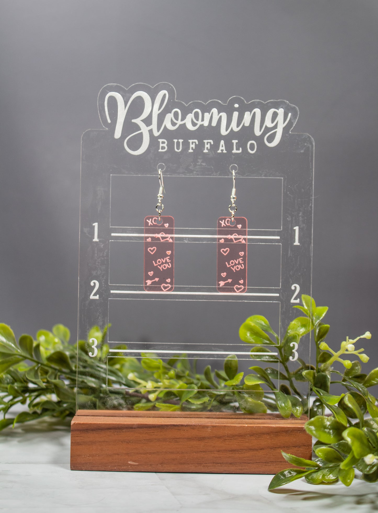 Translucent Valentine's Day Rose Gold rectangle dangle earrings with Love You, XO, and heart drawings engraved. Earrings are on a Blooming Buffalo branded earring stand