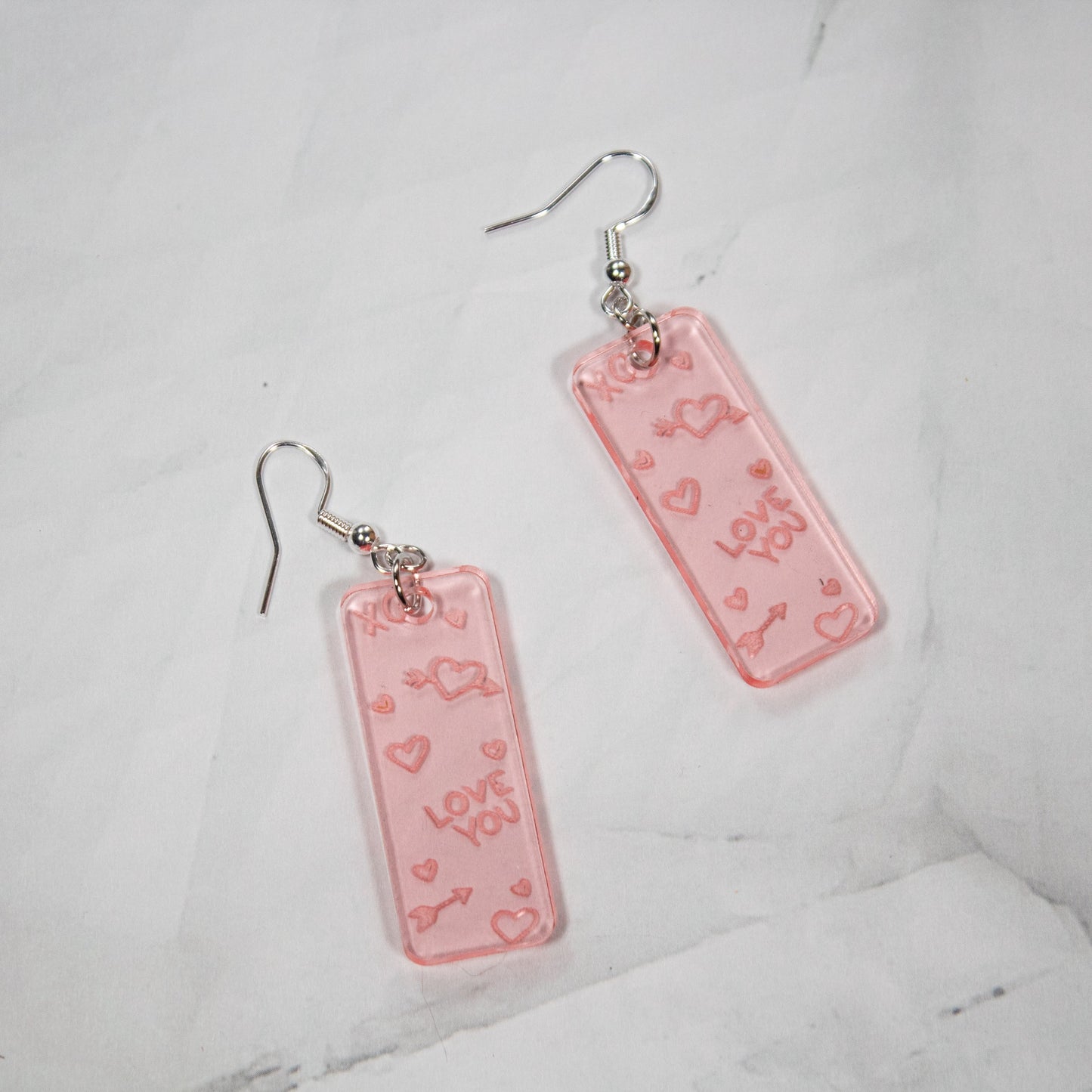 Translucent Valentine's Day Rose Gold rectangle dangle earrings with Love You, XO, and heart drawings engraved. Earrings are on a white marble background