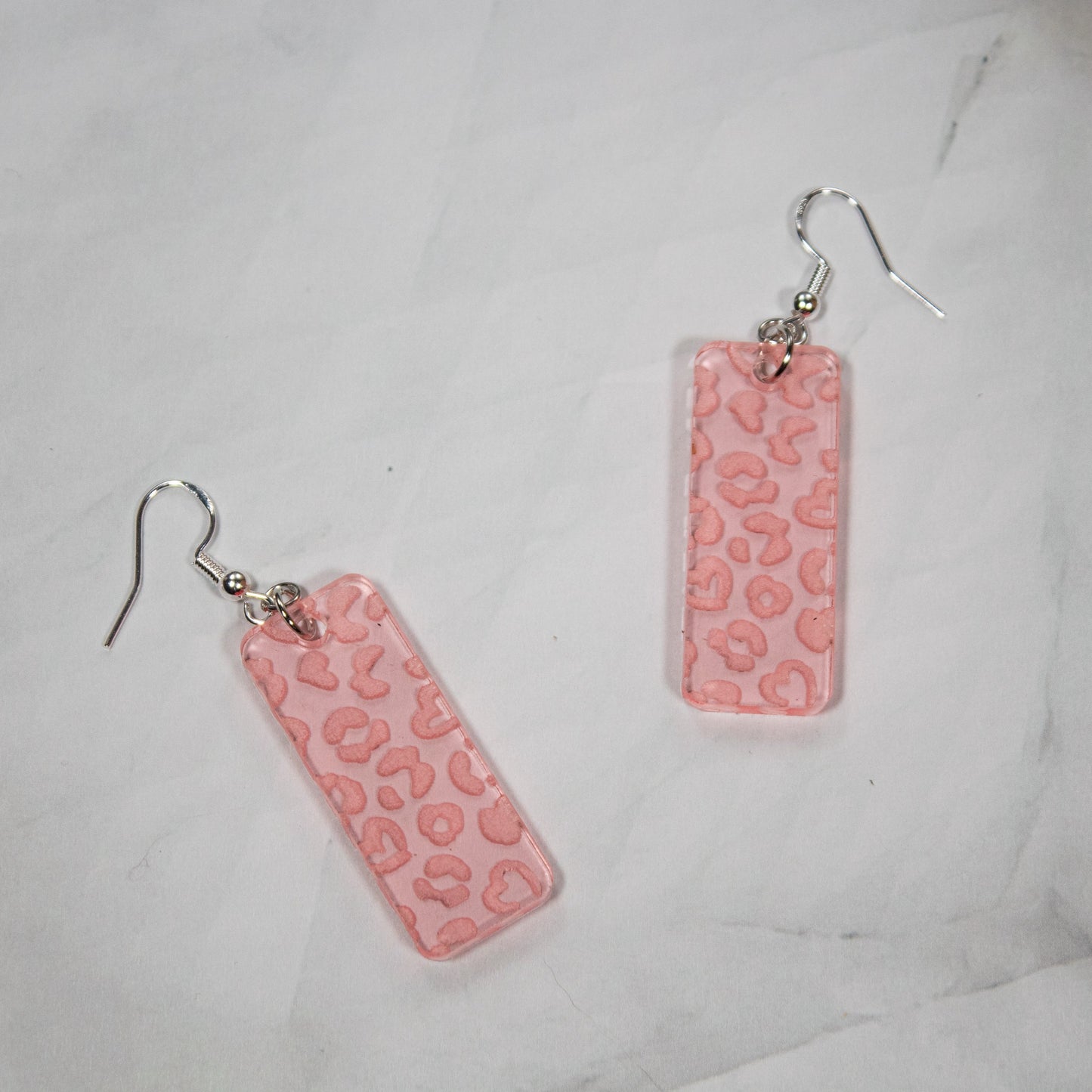 translucent rose gold rectangle acrylic earrings with heart shaped leopard print engraved on earrings. Earrings sitting on white marble background.