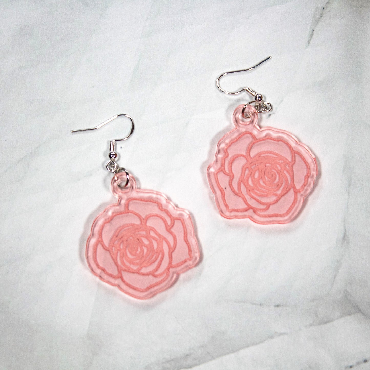 Translucent Rose Gold Acrylic Dangle Earrings shaped like roses with rose detail engraved. Earrings are on a white marbled background.