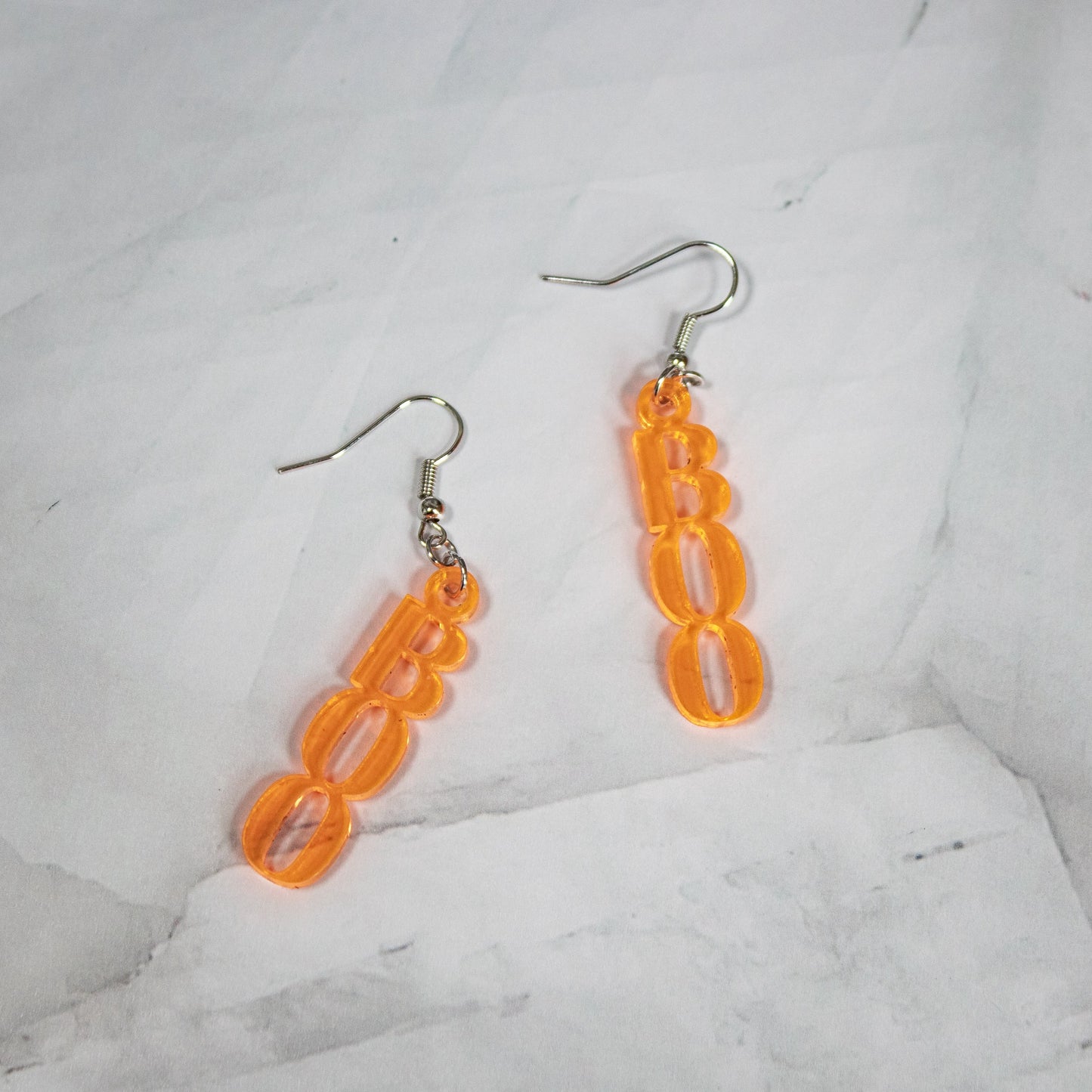 Bright fluorescent orange dangle earrings shaped like the word BOO on a white marble background