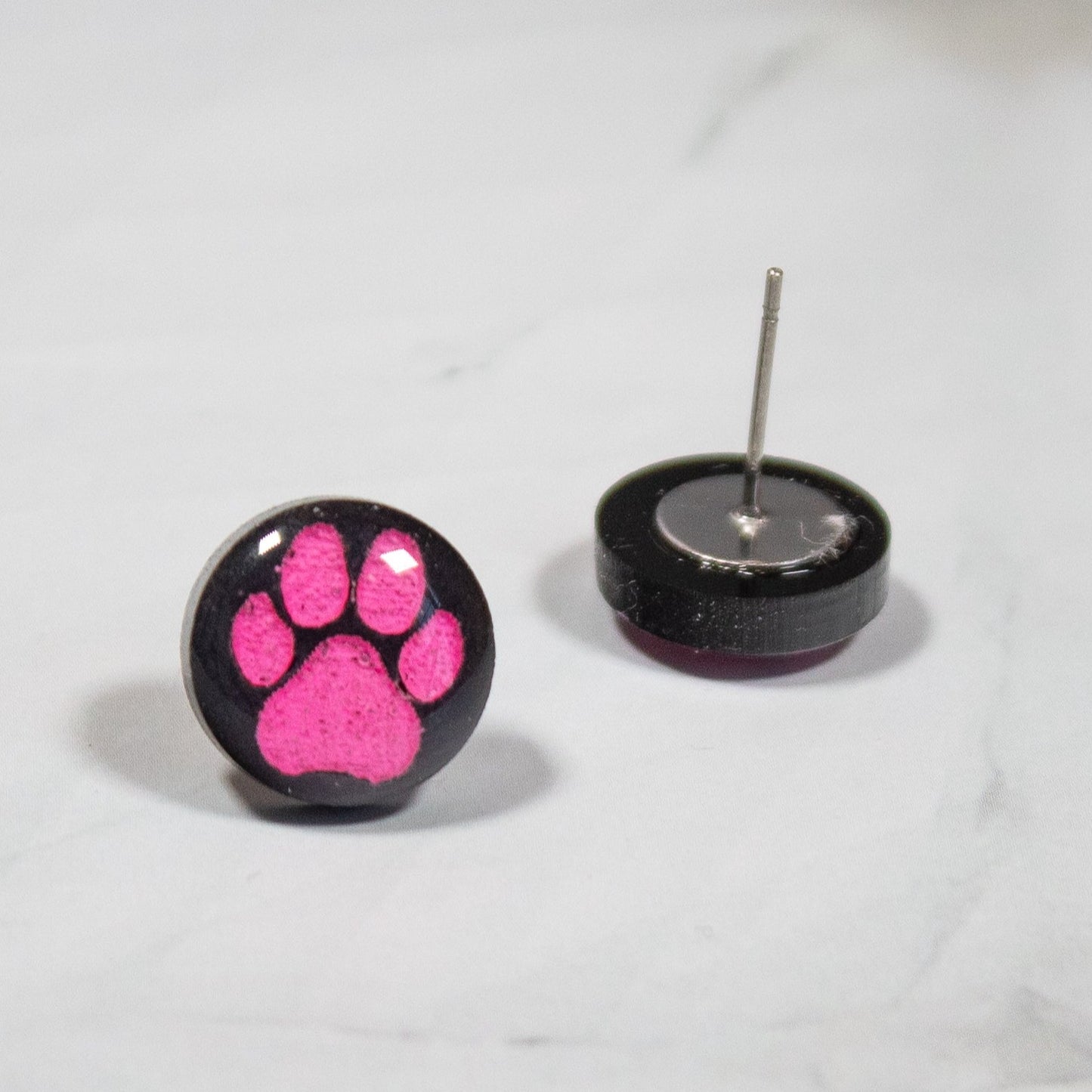 black circular stud earrings with pink pawprints on white marbled background
