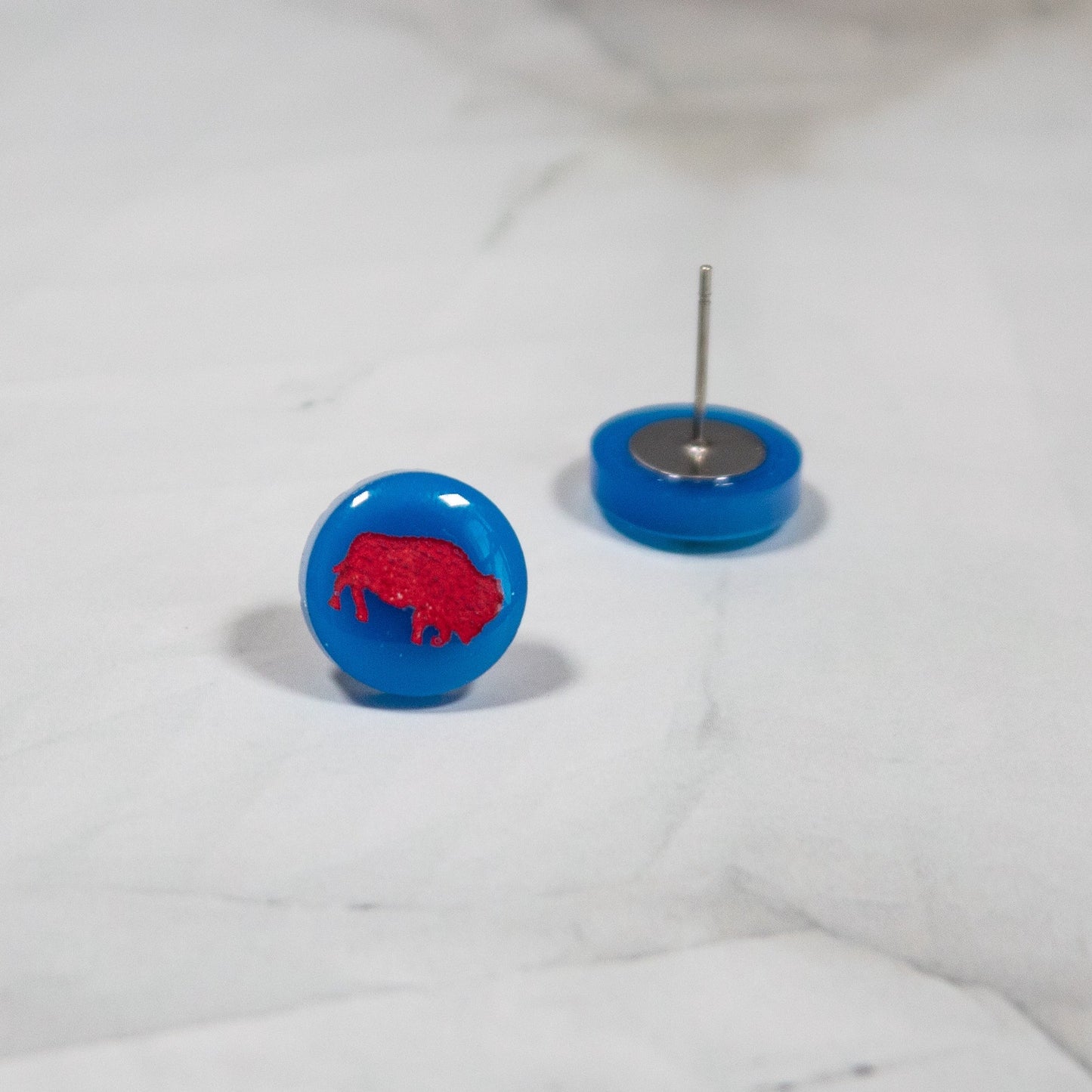 vibrant blue circle stud earrings with a red buffalo design on them. The earrings are resting on a white marbled background