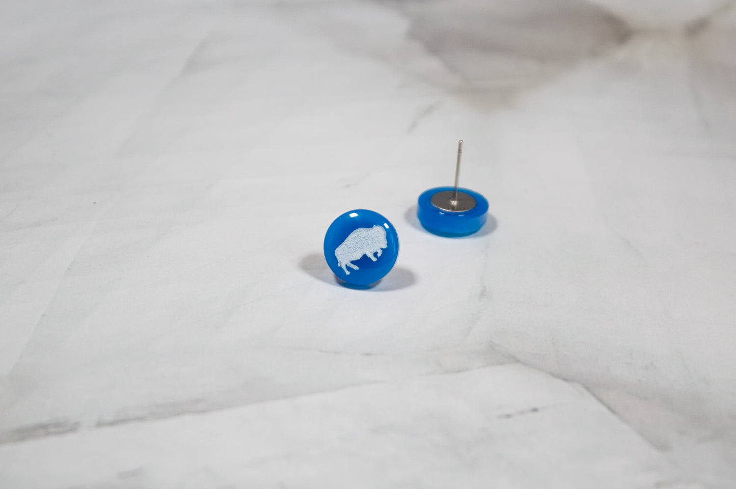 vibrant blue circle stud earrings with a white buffalo design on them. The earrings are resting on a white marbled background