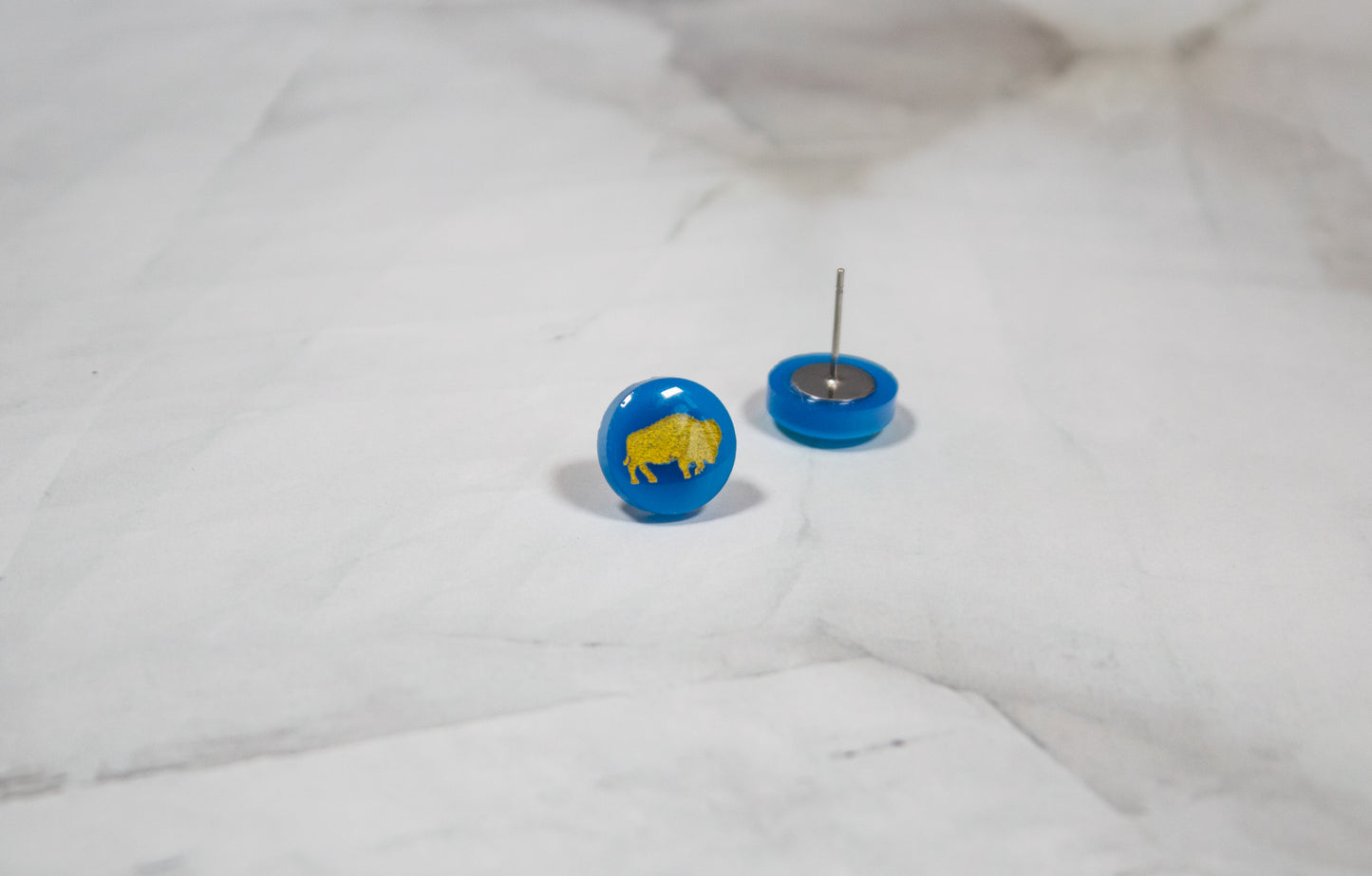 vibrant blue circle stud earrings with a yellow buffalo design on them. The earrings are resting on a white marbled background