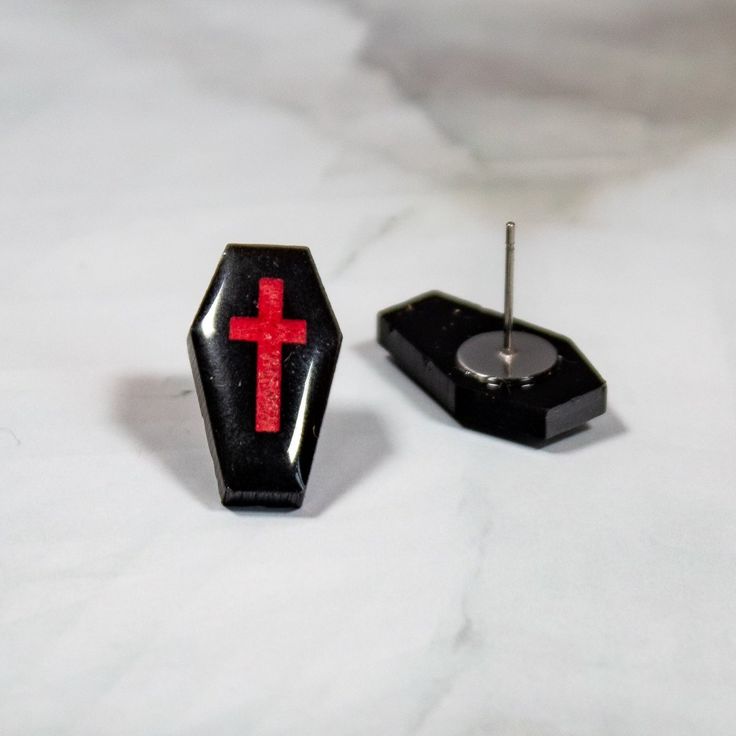 black coffin shaped stud earrings with red crosses on them resting on a white marbled background