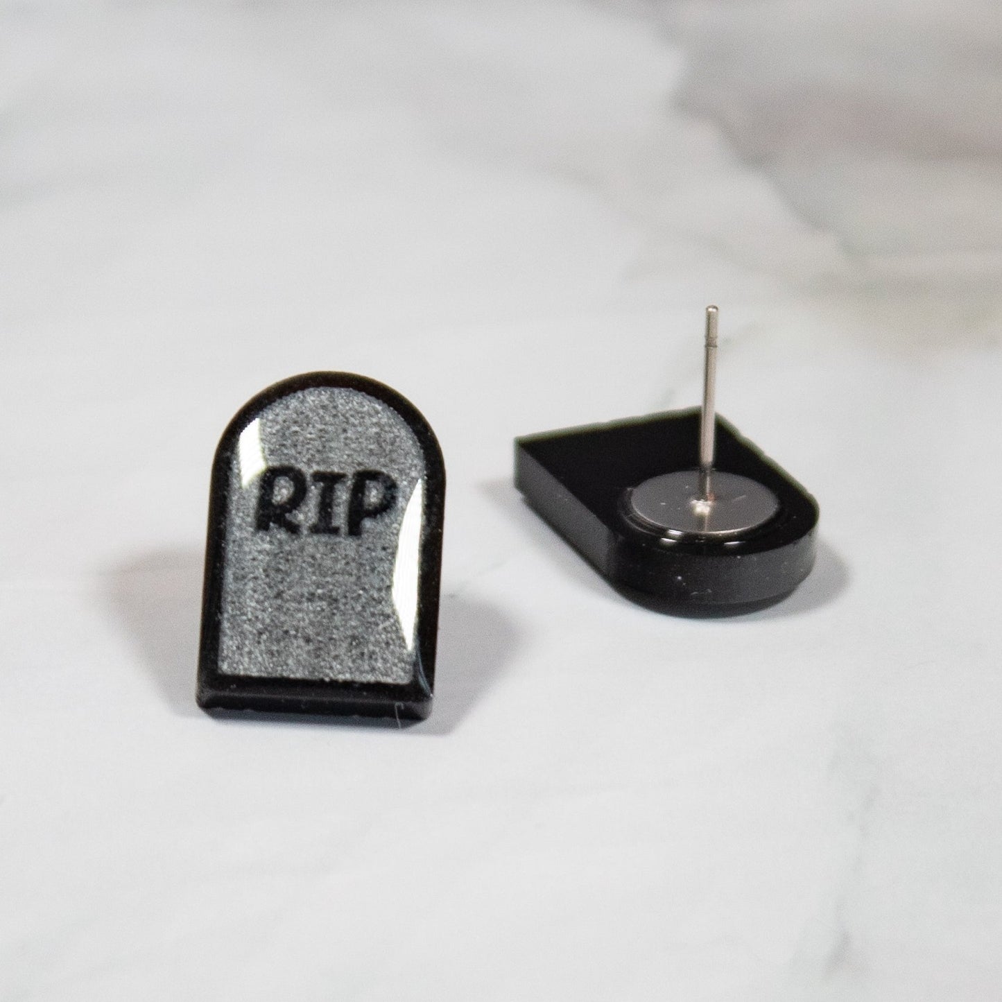 Gray and black tombstone stud earrings with RIP on them resting on white marbled background