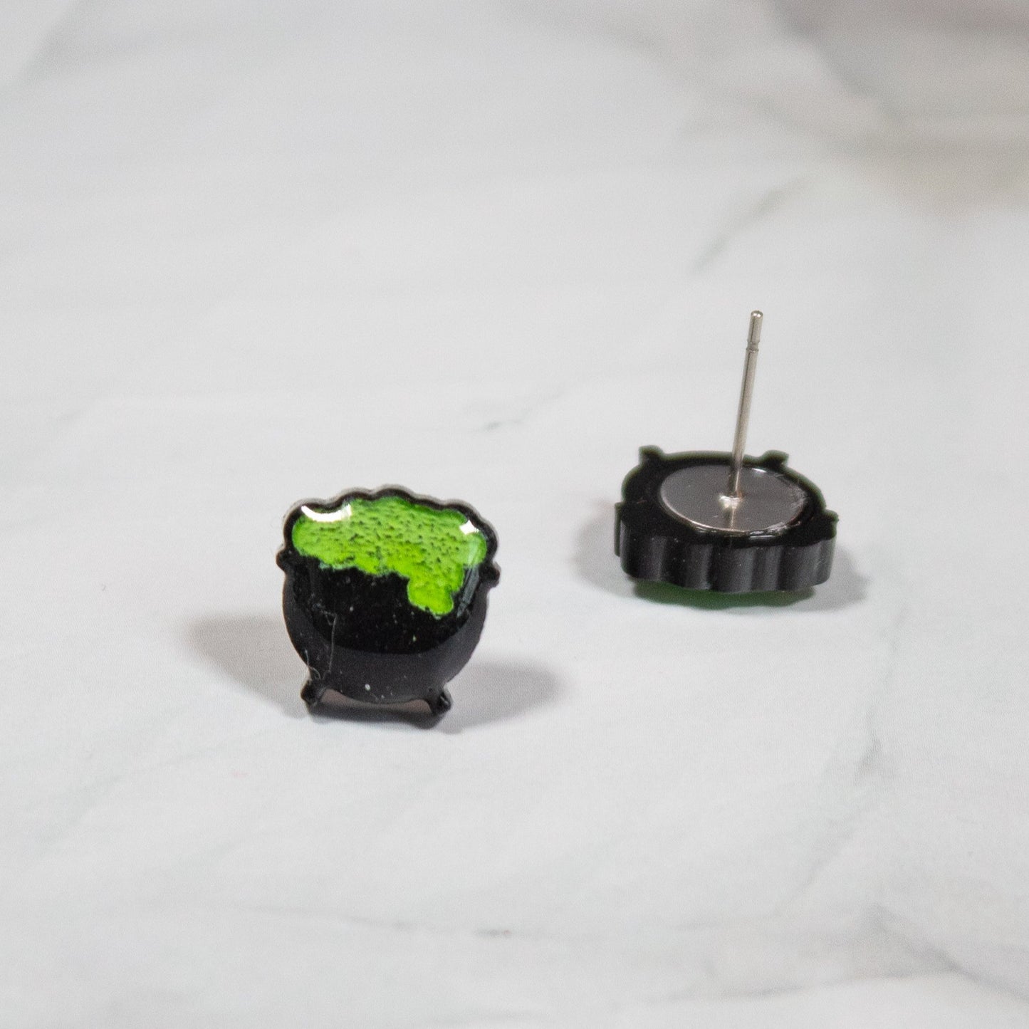 black cauldron stud earrings with vibrant green witch's brew bubbling over the side on a white marble backgound